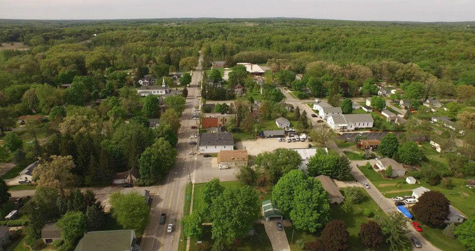 Aerial view of a small town surrounded by lush greenery. The town features residential buildings, a few commercial structures, and tree-lined streets.