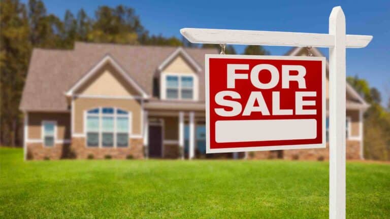 A "For Sale" sign in the foreground with a two-story house blurred in the background, set on a lush green lawn.