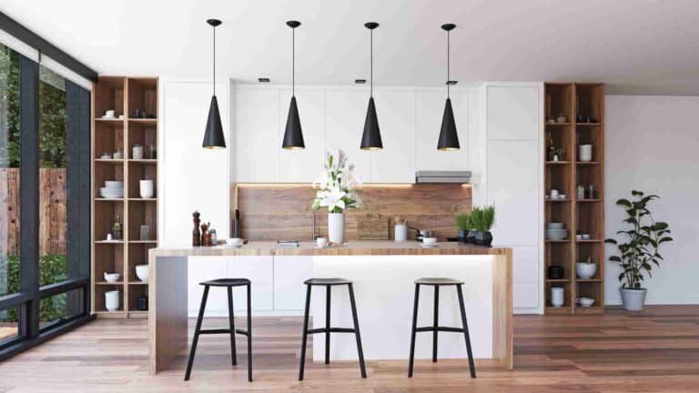 Modern kitchen with white cabinets, black pendant lights, and a wooden island with three black stools. Shelves on both sides hold various kitchenware. Large window on the left provides natural light.