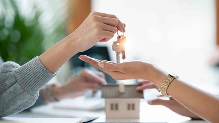 Two people exchange keys over a table with a model house on it.