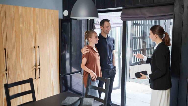 A couple stands in a kitchen with a woman holding a clipboard, likely discussing something related to the home.