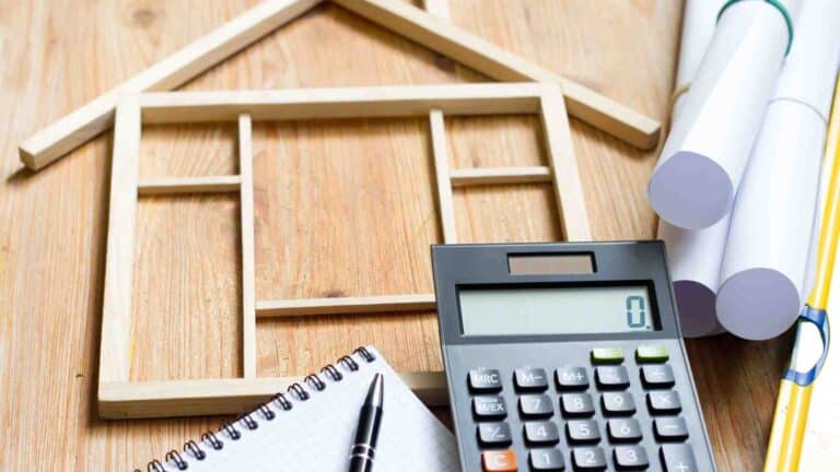 Wooden house frame model, calculator, blueprint rolls, notepad, and pen on a wooden surface.