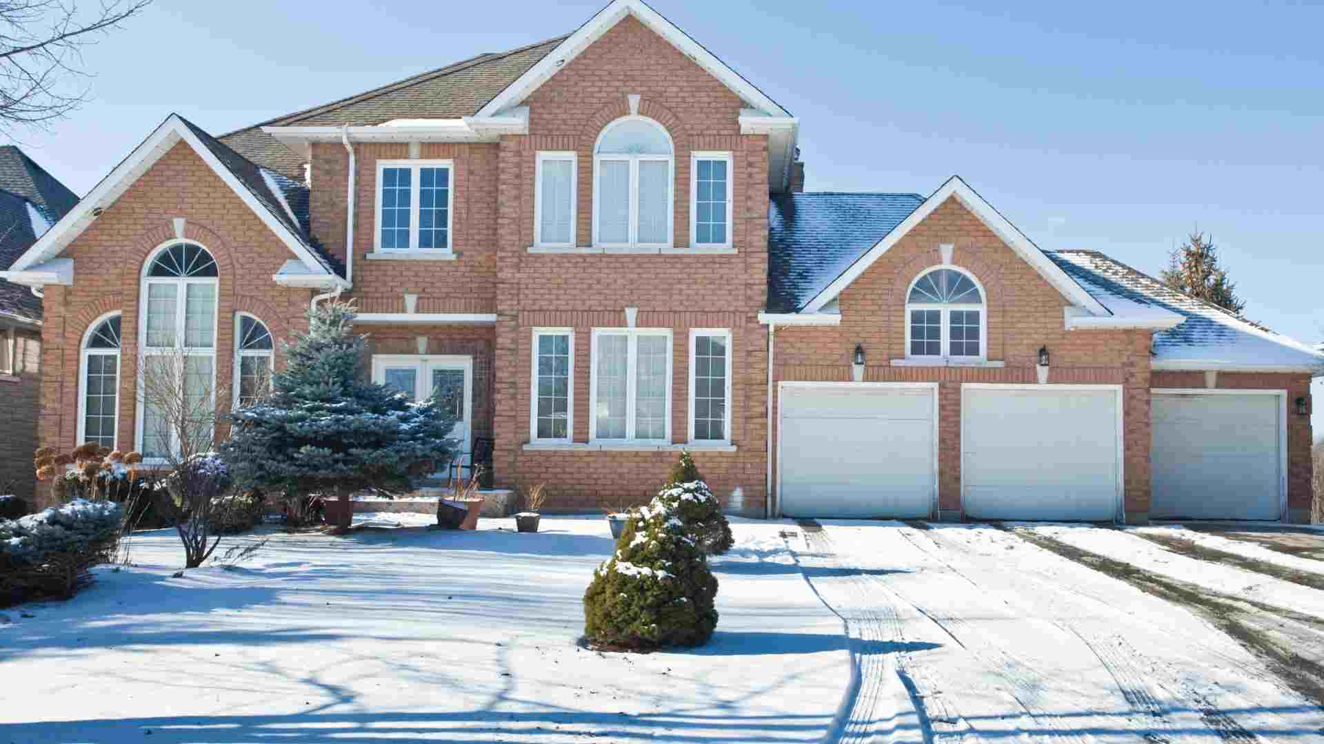 A two-story brick house with three white garage doors and a snow-covered driveway and front yard.