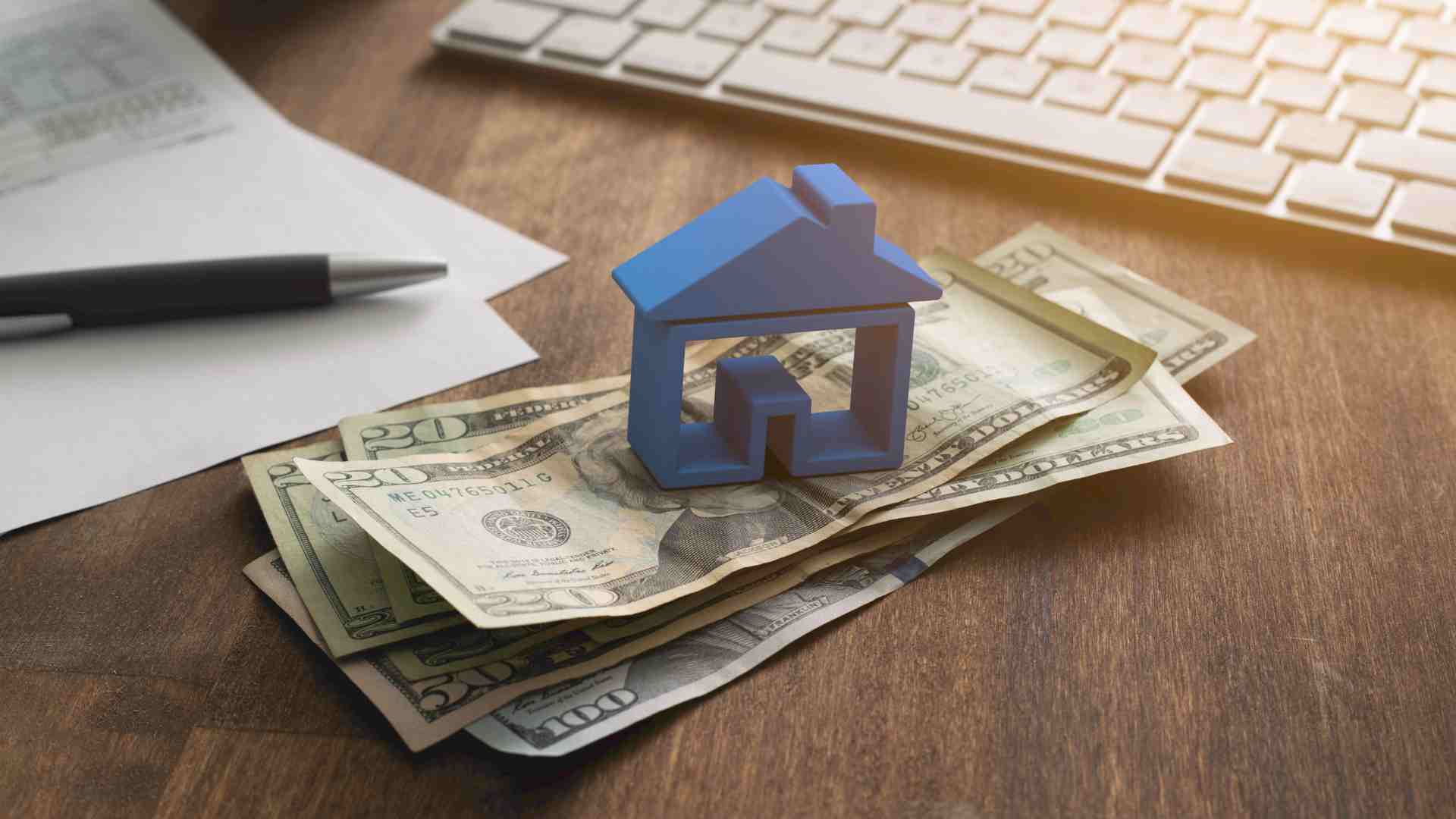 A small blue house model sits atop a stack of US dollar bills on a wooden desk, next to a pen, paper, and computer keyboard.