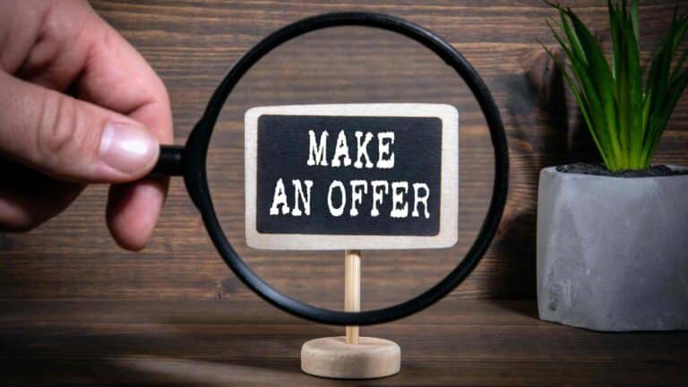 Hand holding a magnifying glass over a small sign that reads "MAKE AN OFFER" against a wooden background with a potted plant.