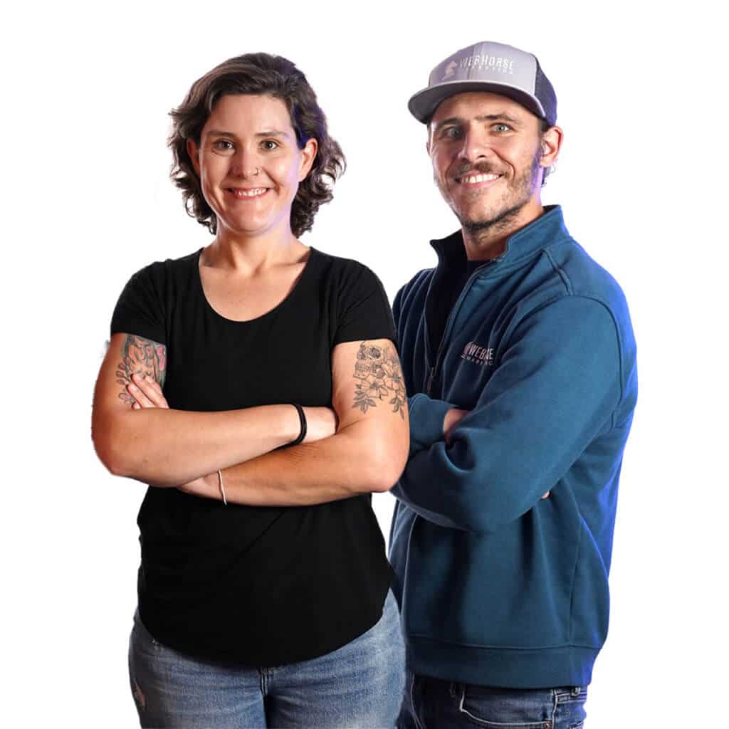 Two individuals standing against a white background. The woman on the left, representing a Michigan real estate team, is wearing a black shirt and jeans, with her arms crossed. The man on the right is wearing a cap and a blue zip-up jacket.